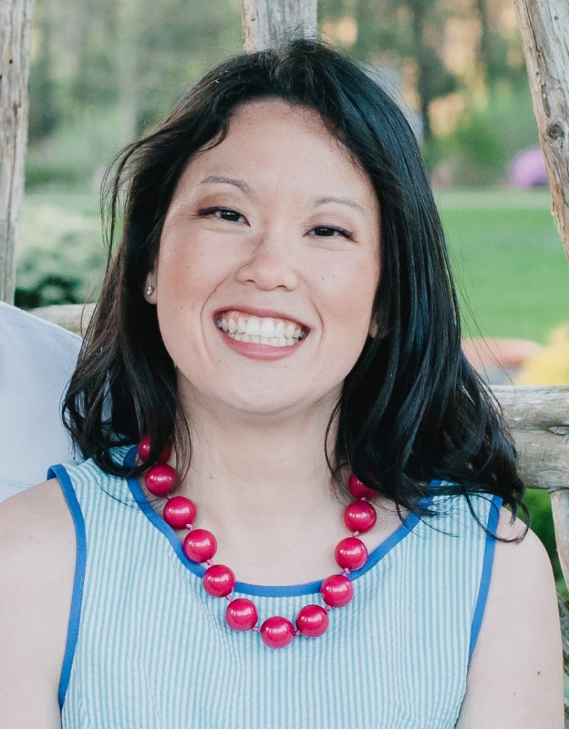 A woman with long black hair is smiling. She wears a light blue and white tank featuring blue piping around the collar & arm openings. Her necklace features large bright red beads. Behind her grass and trees can be seen.
