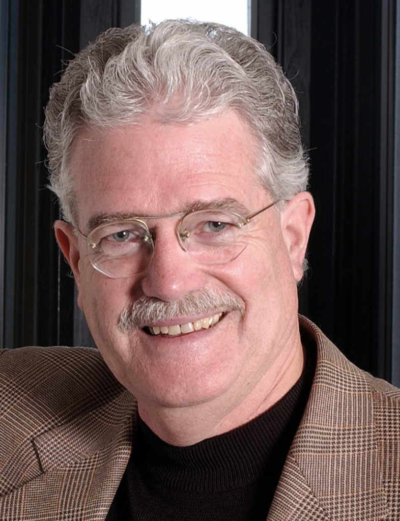 A man with short gray hair is smiling. He has a gray mustache. He wears frameless oval eyeglasses, a black shirt, and a brown glen plaid jacket.