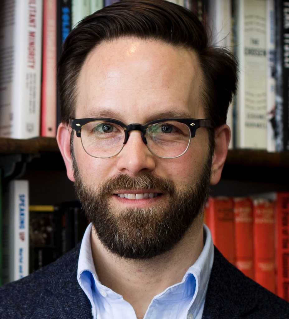 A man with short dark brown hair, mustache, and beard is smiling. He wears black horn-rimmed glasses, a light blue button-down shirt, and a navy tweed jacket. She stands before a bookshelf laden with books.