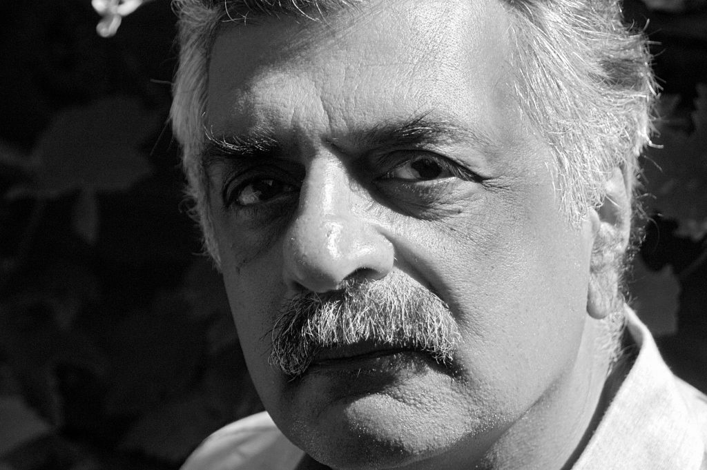 A black and white image of a man with short gray & white hair and mustache. He is bathed in sunlight.