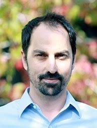 A man stands outside in front of pink, red, and green flora. He has short black hair, mustache, and beard. He wears a light blue button-down shirt.