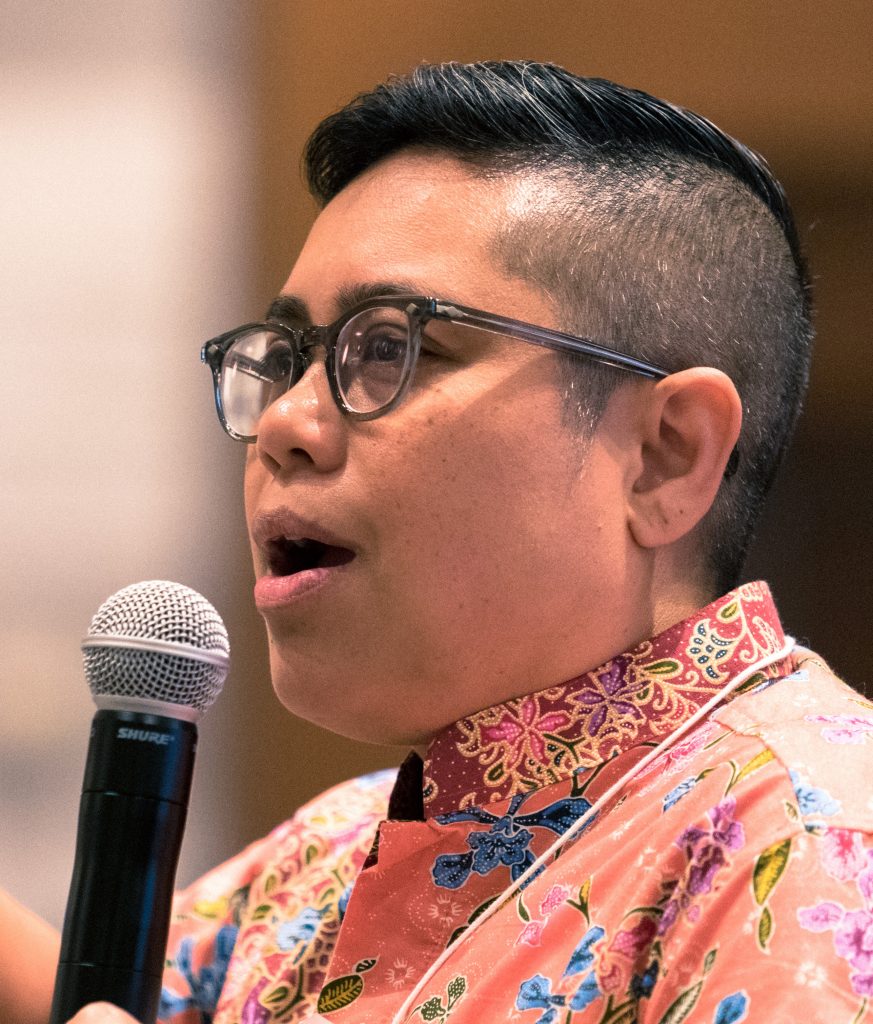 A person holds a microphone and appears to be speaking. They have dark brown hair that is shaved on the sides and back of their head and worn short on the top. They wear dark gray eyeglasses. Their shirt features a bright floral pattern in tones of salmon, blue, green, and purple.