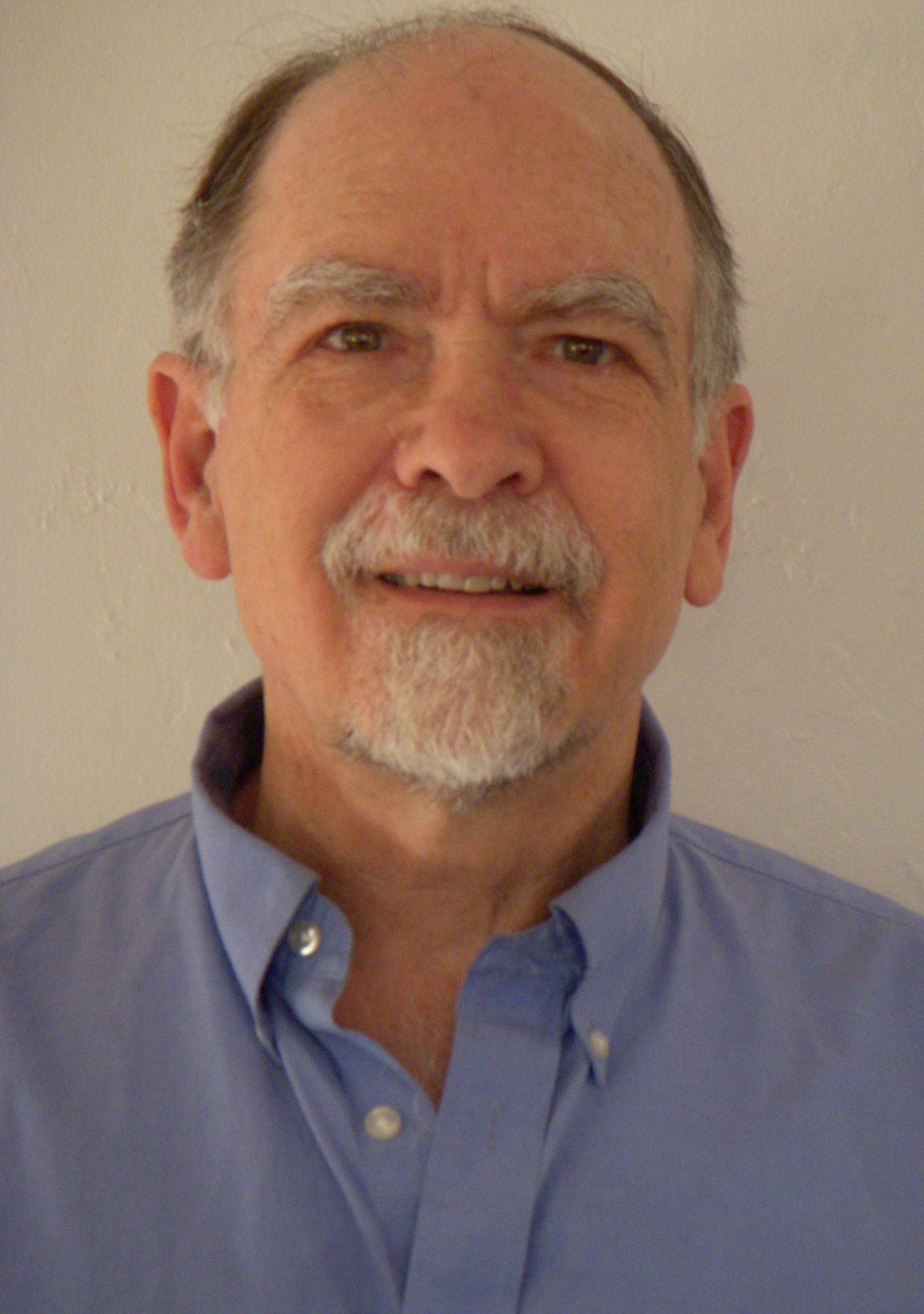 A man with gray hair, white mustache, and white goatee is smiling. He wears a light blue button-down shirt.
