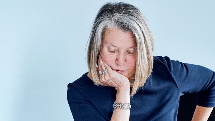 A woman with a blond shoulder-length bob rests her head on her right hand and gazes downwards. She wears silver and stone rings on her right hand, as well as a looped silver-toned bracelet. She wears a navy blue boatneck top, and her left arm appears to be perched on the back of a chair not in frame. She sits before a blue background.