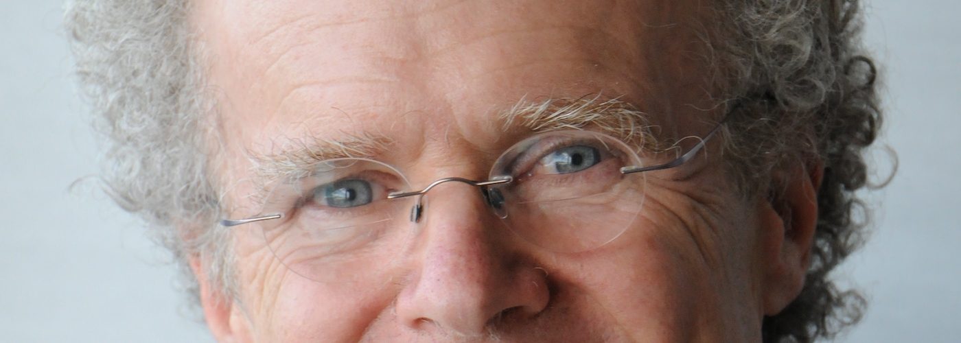 A man wearing oval frameless eyeglasses is smiling. He has short curly white hair. He wears a purple shirt and a brown leather vest.