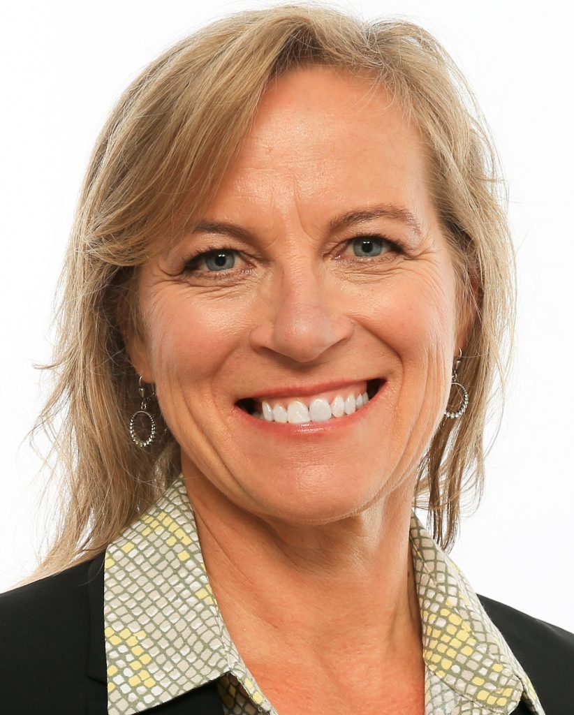 A smiling woman with shoulder-length blond hair. She wears earrings with circular drops, a patterned blouse in tones of yellow and gray, and a black suit jacket.