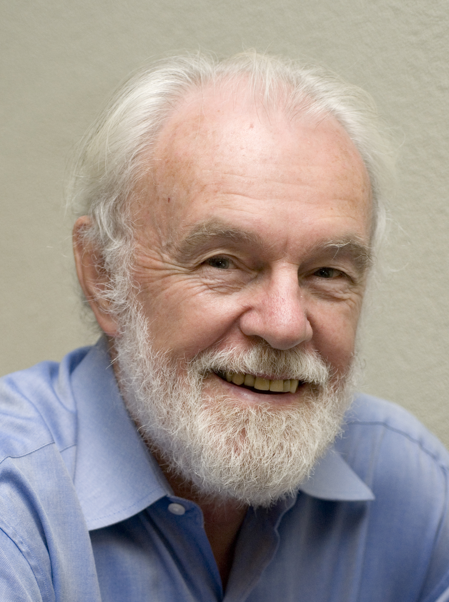 A smiling man with white hair, mustache & beard wears a light blue button-down shirt. He poses against a white wall.