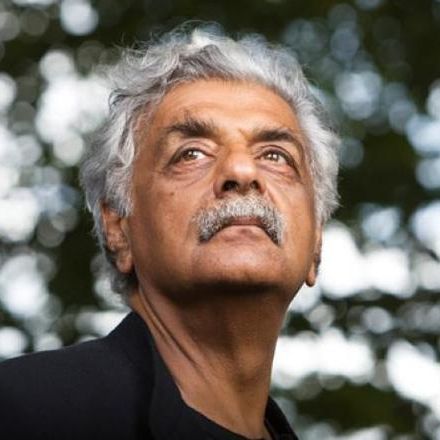 A man with white hair and mustache looks out into the distance. He wears a black shirt and jacket. In the background foliage can be seen.