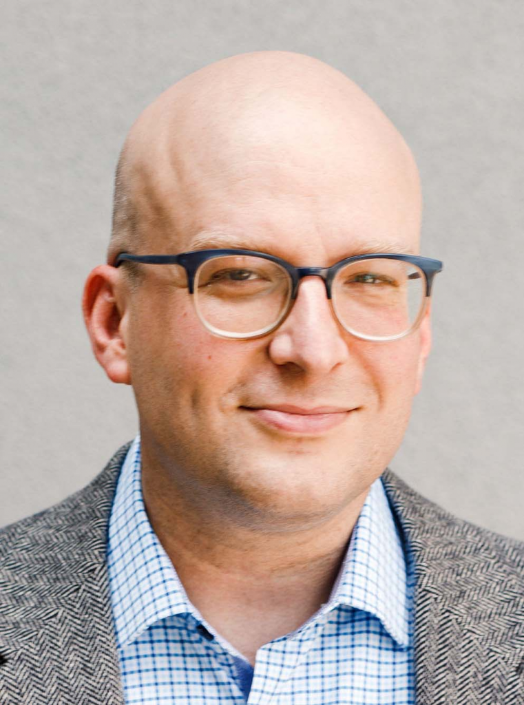 A bald man wearing two-toned eyeglasses smiles. He is wearing a blue and white windowpane button-down shirt and a black and white herringbone jacket.