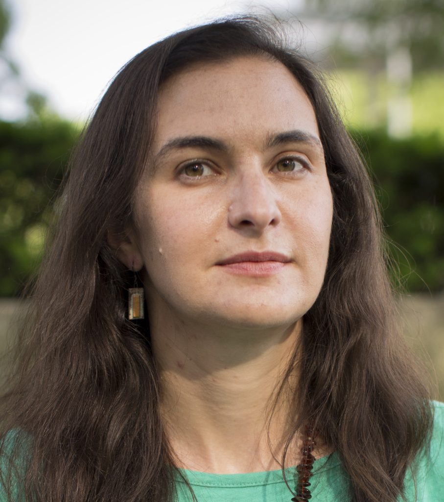 Woman with long dark brown hair wearing bright green shirt, drop earrings, and brown stone necklace. Greenery can be seen behind her.