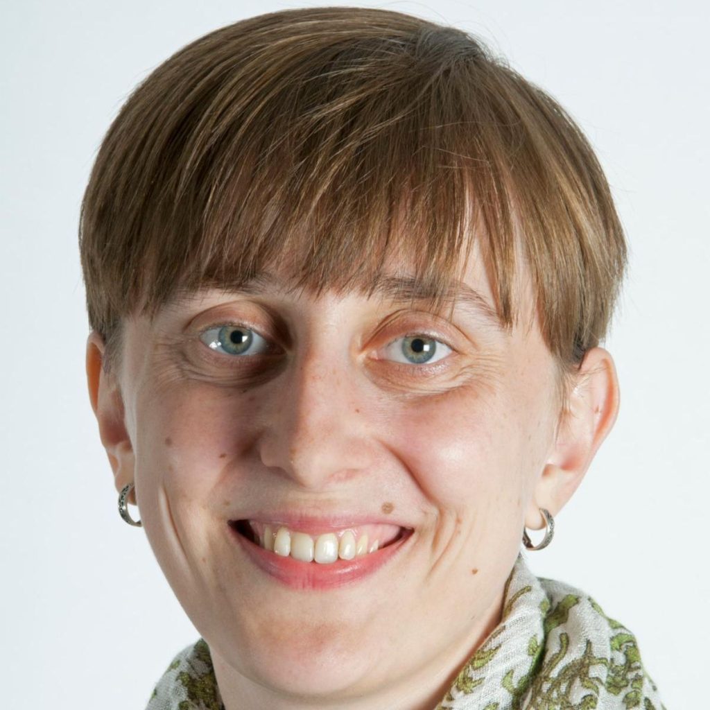 Smiling woman with short brown hair wearing small silver hoop earrings and a green & white scarf.