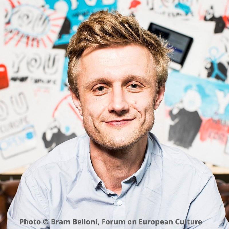 Smiling man with blond hair wearing a light blue button-down shirt. A collage of images in red, blue, black, and white can been seen in the background.