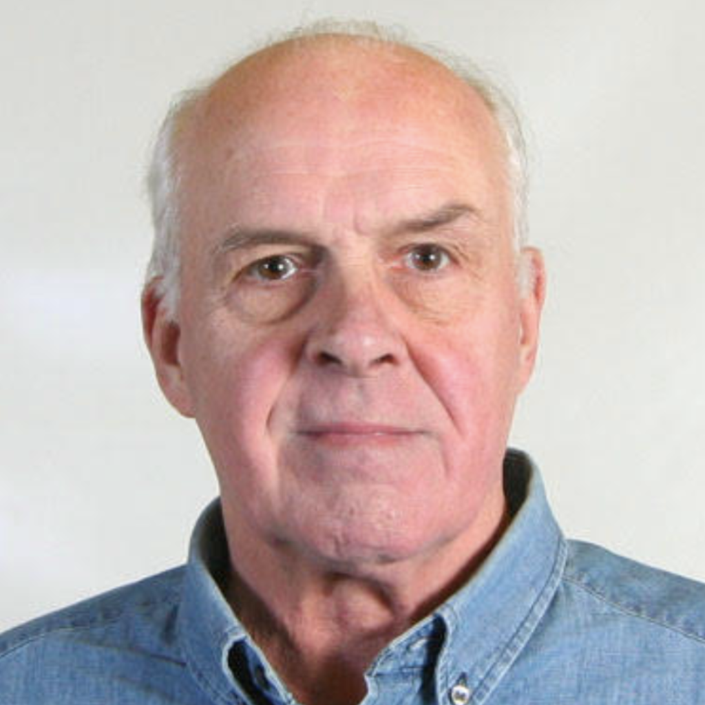 A man with white hair smiles slightly wearing a chambray button-down shirt.