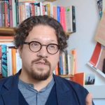 A man with short, curly dark brown hair, mustache & goatee sits in front of a colorful array of books. He wears round eyeglasses, a dark gray henley, navy blue cardigan, and navy blue jacket.