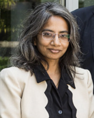 A women with medium length hair and glasses looks at the camera. She has a slight smile and is wearing a black dress shirt with a beige suit jacket.