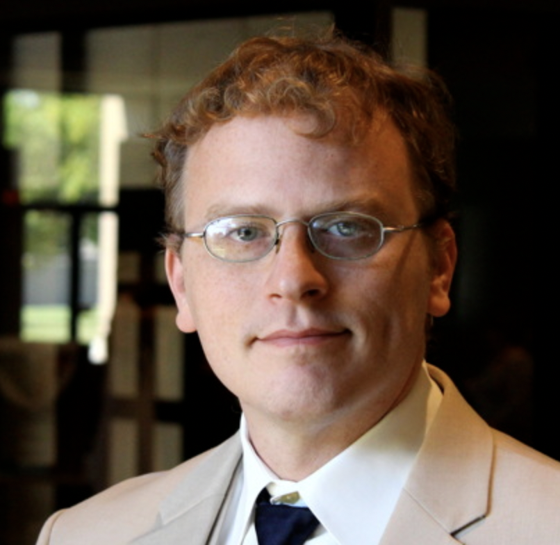 A man with glasses and short curly light brown hair looks straight at the camera. He is wearing a light colored suit and dark blue tie. He is inside a building and behind him is a sunlit window.