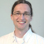 A man is smiling broadly at the camera. His long, light brown hair is parted down the middle and gathered behind his head in a low ponytail. He wears metal eyeglasses featuring oval lenses that appear frameless on the lower halves. He wears a white button-down shirt and white undershirt. He stands before a light-blue backdrop.