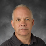 A man stares intently into the camera. He is seated before a dark gray background. His white and gray hair is worn short, and he has blue eyes. He wears a dark gray button-down shirt.