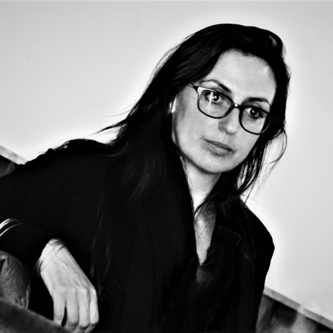 A black and white photo of a woman with dark mid-length hair and glasses looking to the left of the camera into the distance. Her elbow is bent and we can see her right hand resting on the seat. The background is white.