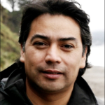 A man looks directly at the camera, his mouth slightly open as if caught about to smile. His short black hair appears blown back from his face, suggesting a breeze. He wears a black waterproof parka. He is outside. A green hillside with a tree can be seen behind his right shoulder, and over his left, a beach, a body of water, and a distant shoreline. The color of the sky is white, like the water.