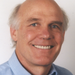 A man smiles broadly at the camera. His white hair is worn short, and the top of this head is bald. He wears a light blue button-down shirt. A white wall is seen in the background.
