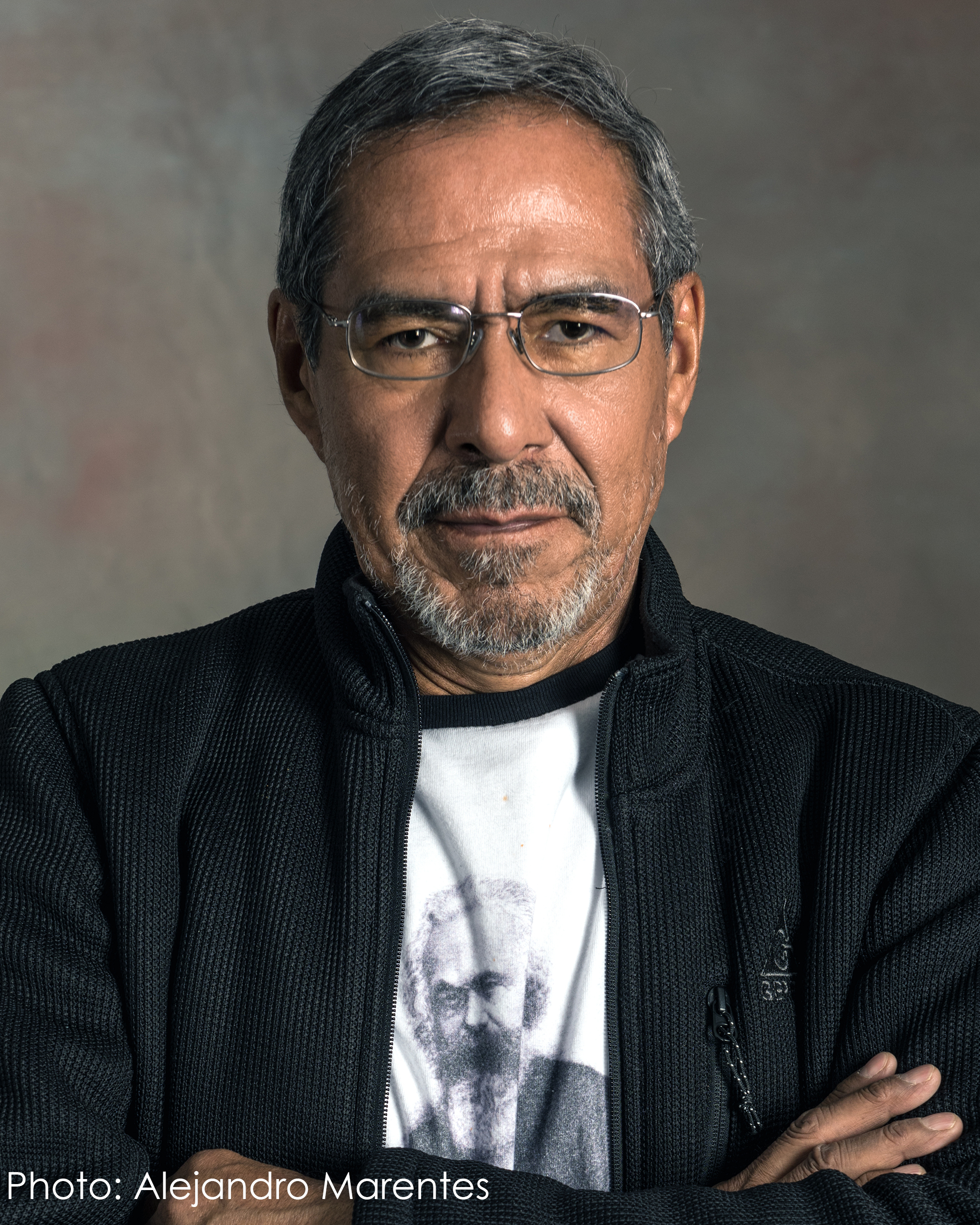 A man looks confidently into the camera. His arms are crossed in front of his chest. His short, black hair is peppered by shades of white and gray. His mustache and beard, featuring similar coloring, is worn close to his face. His eyeglasses, in a silver metallic tone, feature rectangular lenses. He wears an unzipped black woven athleisure jacket with a stand-up collar. A vertical pocket seam with a black-and-white woven pull cord is seen on his left chest. Beneath the jacket, he wears a bright white t-shirt with a black-ringed color, which features the black-and-white image of Karl Marx. He stands before a brown-gray backdrop.