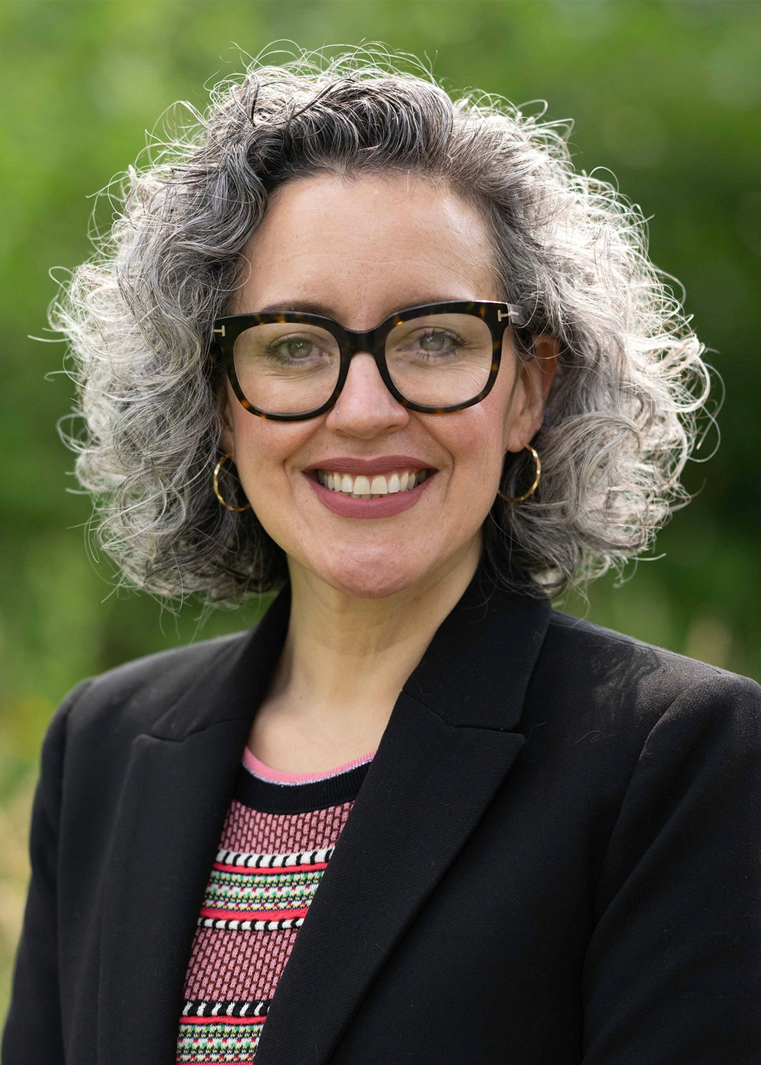 A woman stands before lush green foliage. She smiles broadly as she looks directly into the camera. Her curly, salt-and-pepper hair is worn in a chin-length bob with an offside part. Her oversized, dark tortoiseshell eyeglasses feature wide, rectangular lenses. She wears a black wool blazer atop a crewneck sweater that features stripes and patterns in shades of pink, white, green, and black. Medium-sized gold hoop earrings hang from her ears.
