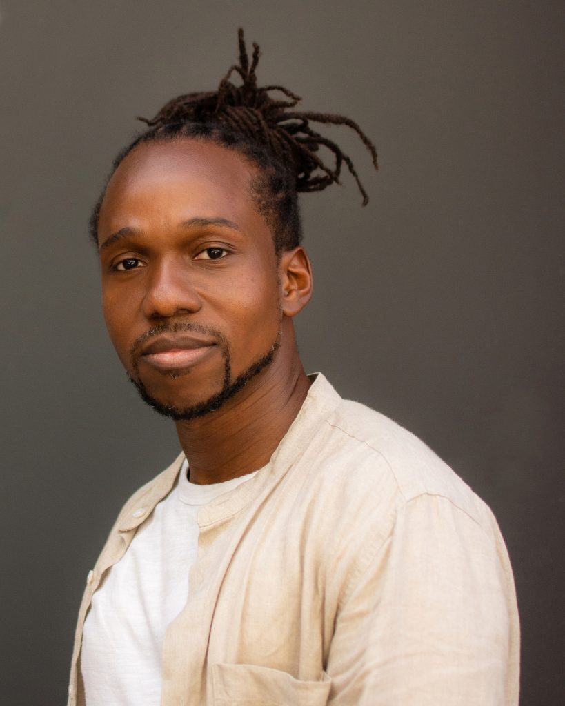 A man smiles warmly at the camera before a dark gray backdrop. His dark-brown hair, which has been plaited into braids, is gathered atop the crown of his head in a messy bun. He wears his facial hair - mustache and beard - close to his face. He wears a cream button-down shirt that is undone to reveal a white t-shirt beneath.