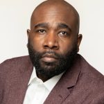 A man looks directly into the camera positioned before a white backdrop. His hair is shorn so closely to his scalp that he appears bald. His black mustache and beard are also shorn close to his skin. He wears a crisp white button-down shirt and a mauve tweed suit jacket.