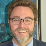 A man stands before a vibrant blue-green print featuring black lines which may be branches of a plant or flower. He is smiling directly at the camera. His light brown hair is short and worn off his face. His light brown mustache and beard is also worn short and peppered with gray. He wears a light blue button-down shirt with a black knitted tie. He also wears a dark gray jacket. His acetate eyeglasses are a blue-gray color and feature lenses in an inverted trapezoidal shape.