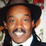 A man smiles broadly at the camera. His braided, dark black hair peeks out beneath a black fedora hat with a wide satin ribbon. He has a black mustache. He wears a bright white button-down shirt, a black-striped satin tie, and a black suit jacket. He stands in front of a wall featuring floral wallpaper, a fire alarm sensor just visible above his head.