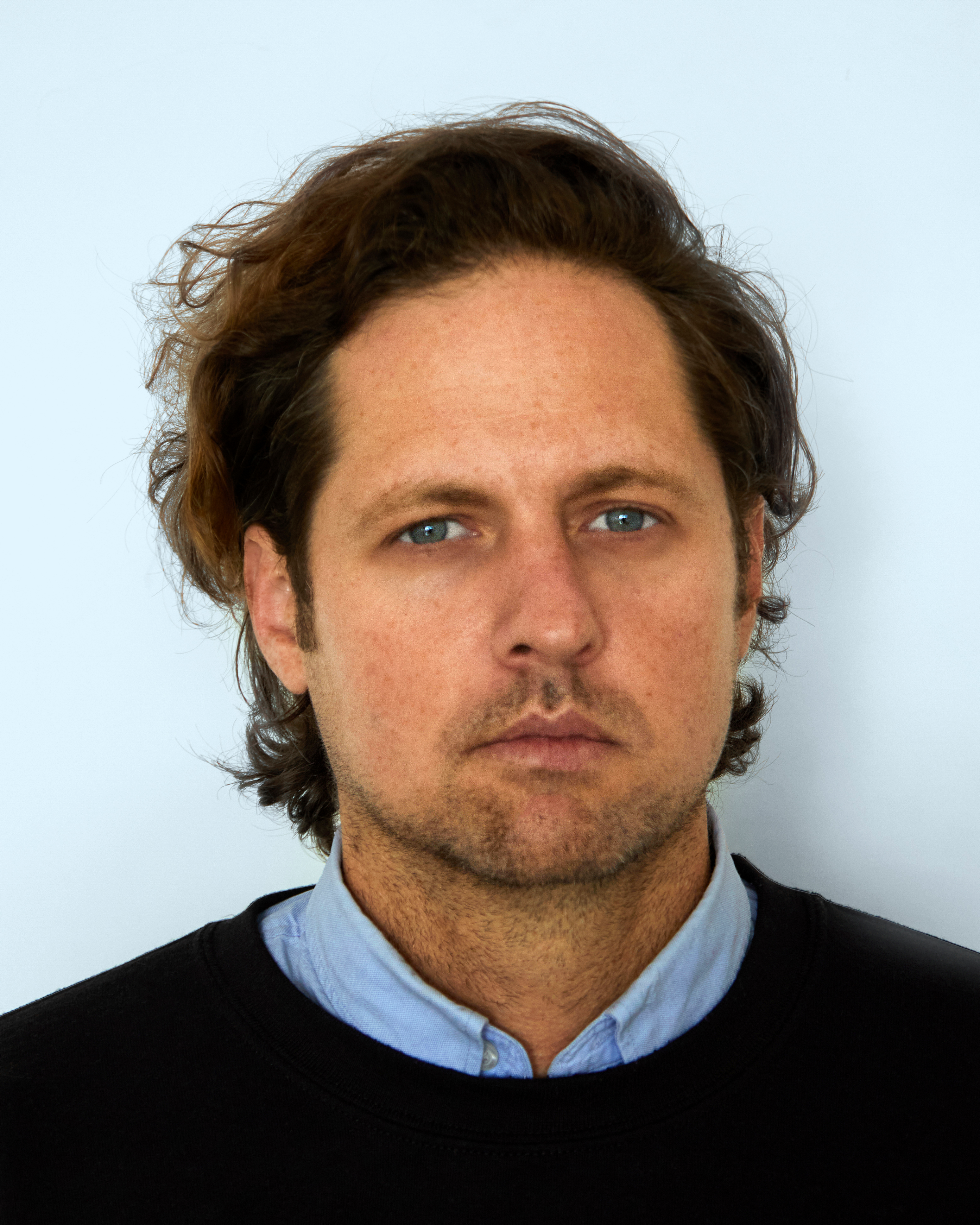 A man looks directly at the camera. He is positioned before a white wall. His dark brown hair is curly and brushed away from his face, which is covered in stubble. He wears a light-blue button down shirt beneath a black crewneck sweater.