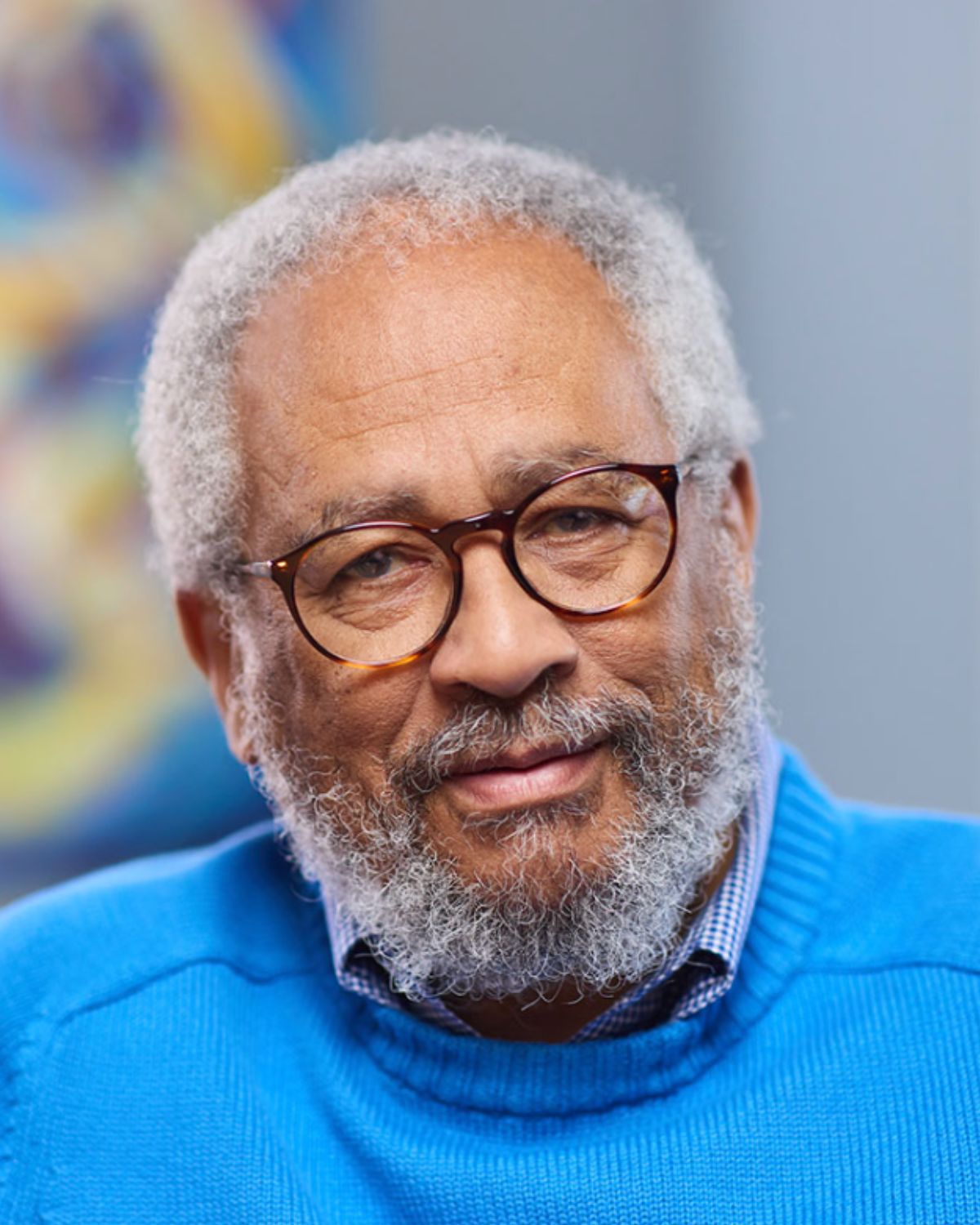A man looks into the camera. His gray and white curly hair is worn short, as is his mustache and full beard. His tortoise-shell eyeglasses have rounded rectangular lenses. He wears a bright blue sweater atop a gingham button-down shirt. Behind him, a colorful piece of abstract wall art can be seen.