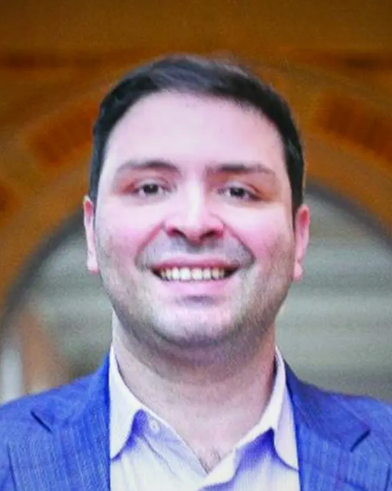 A man stands before an orange-hued archway smiling broadly. He has short black hair, and stubble covers his face. He wears a light blue oxford shirt beneath a cadet-blue suit jacket.