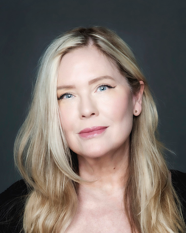 A woman looks directly into the camera, her head at a slight tilt. Her long blond hair is worn in layers that frame her face. She has tucked some behind her ear to reveal a small metallic stud. She wears a black velvet shirt. She sits before a dark gray background.