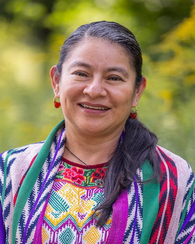 A woman smiles broadly at the camera. Her long black hair, peppered with bits of white, is parted and pulled back into a low ponytail, which hangs over her shoulder. She wears a shirt and shawl that feature various patterns, like arrows, diamonds, birds, and roses, in colors pink, purple, yellow, and green. She wears hoop earrings that feature a red design. Behind her, one sees the suggestion of vibrant greenery.