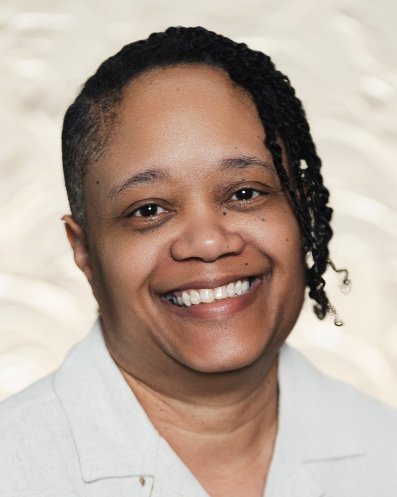 A person smiles broadly at the camera. Their dark brown hair is shorn tight on one side of their head, while the rest is worn in short braids. They wear a light gray collared shirt. Behind them, a soft ivory background can be seen.