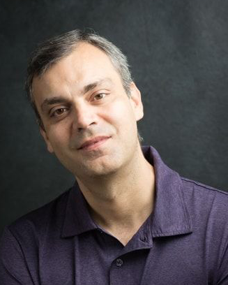 Man with dark hair wearing a dark purple shirt. He is standing in front of a dark background.